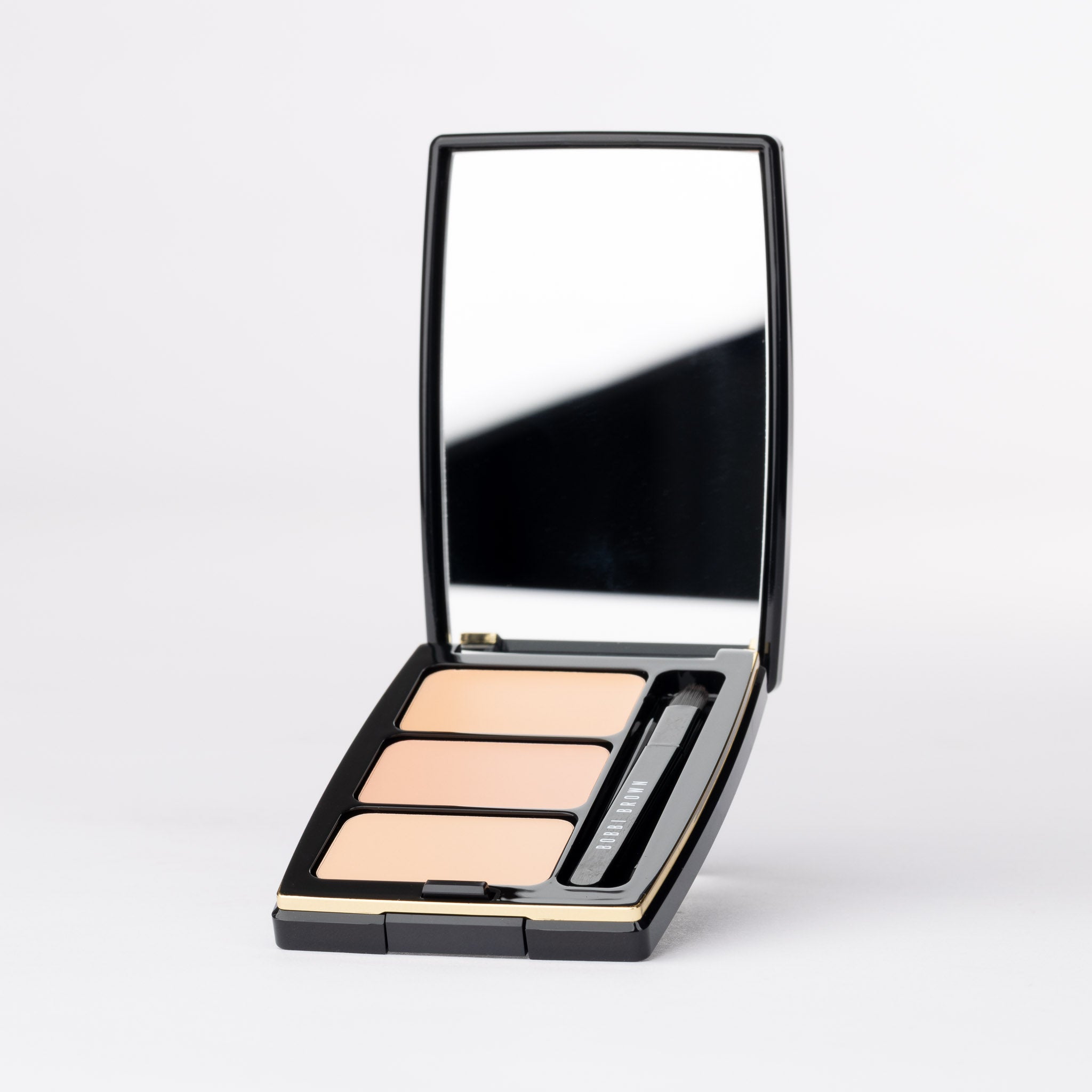 Bobbi Brown luxury black compact palette, open. Mirror, brush & three light beige/peach complexion makeup.